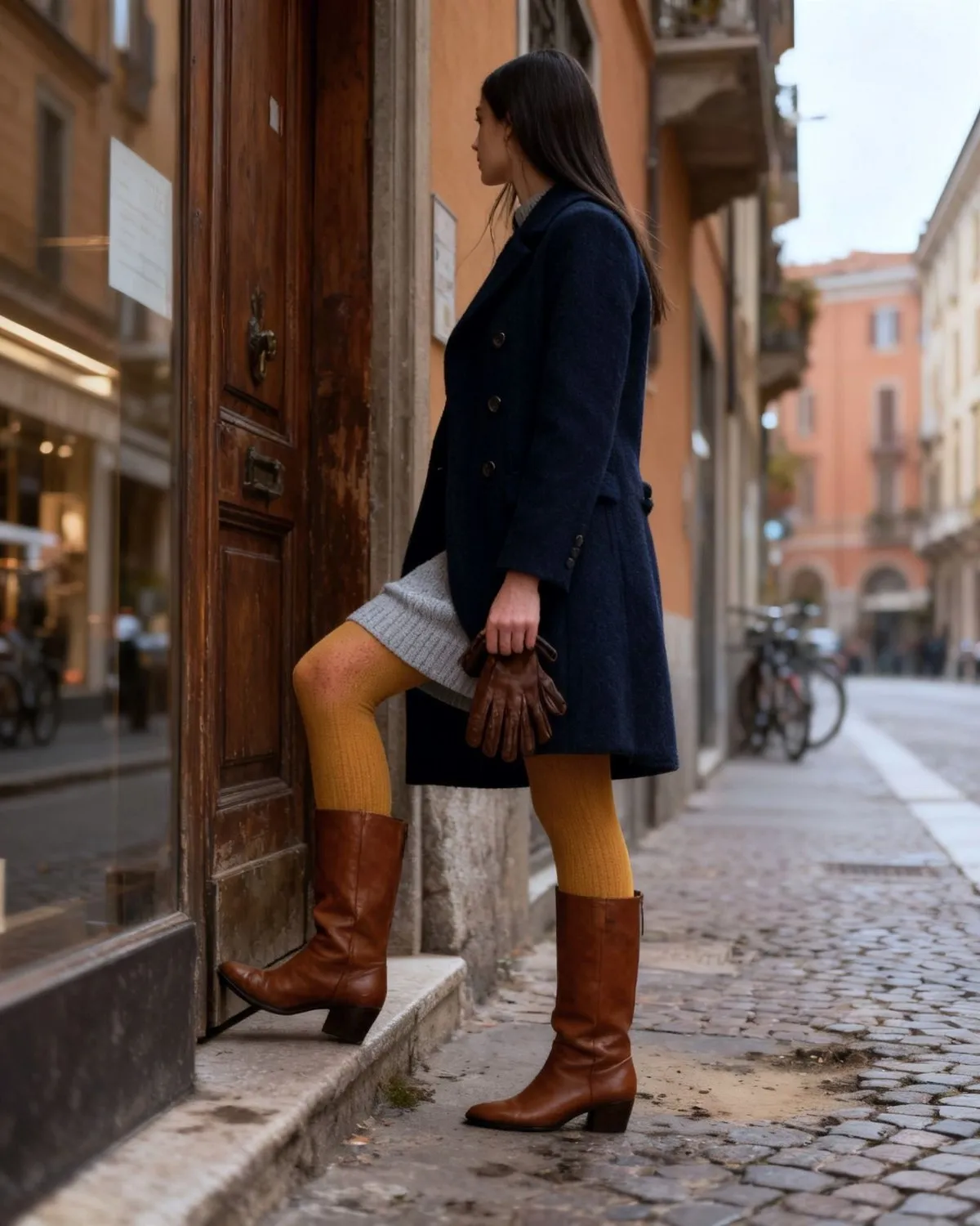 Ava davanti a un portone di legno a Milano, girata verso la strada, con cappotto blu notte doppiopetto, abito in maglia grigio perla, collant senape e stivali al ginocchio color cuoio