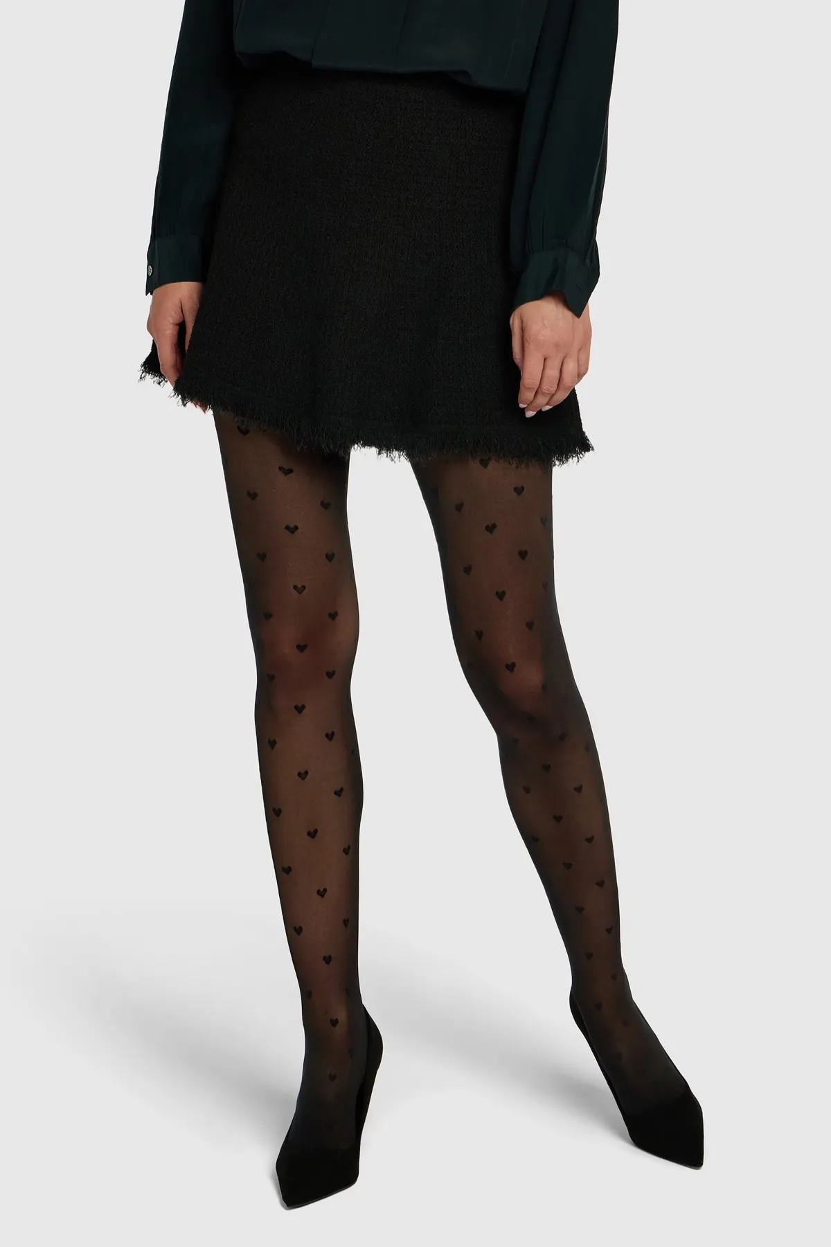 Gambe in collant neri velati con piccoli cuori lavorati, sotto una minigonna di tweed nera, con décolleté nere a punta