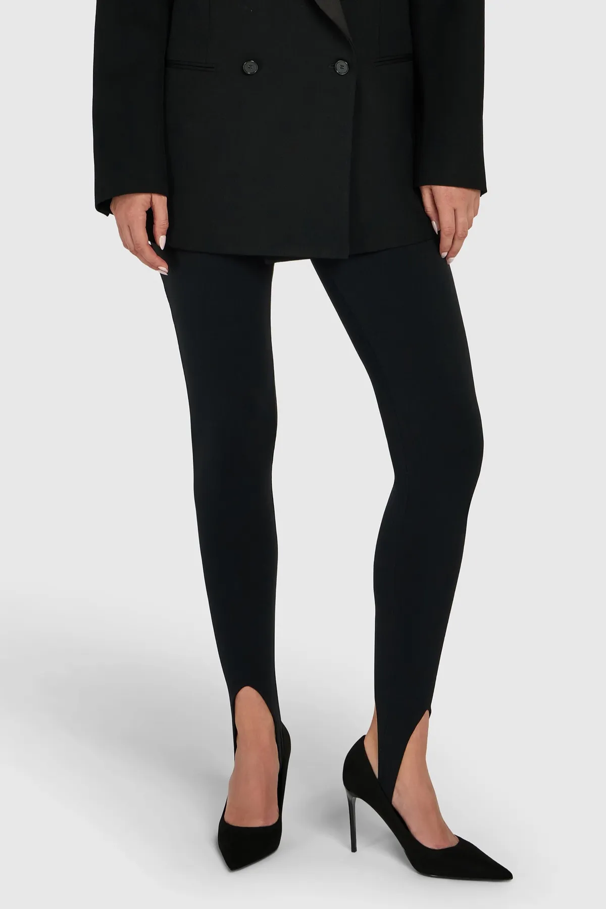 Legs in opaque black stirrup leggings under a black double-breasted jacket, with the stirrup visible over black pointed pumps