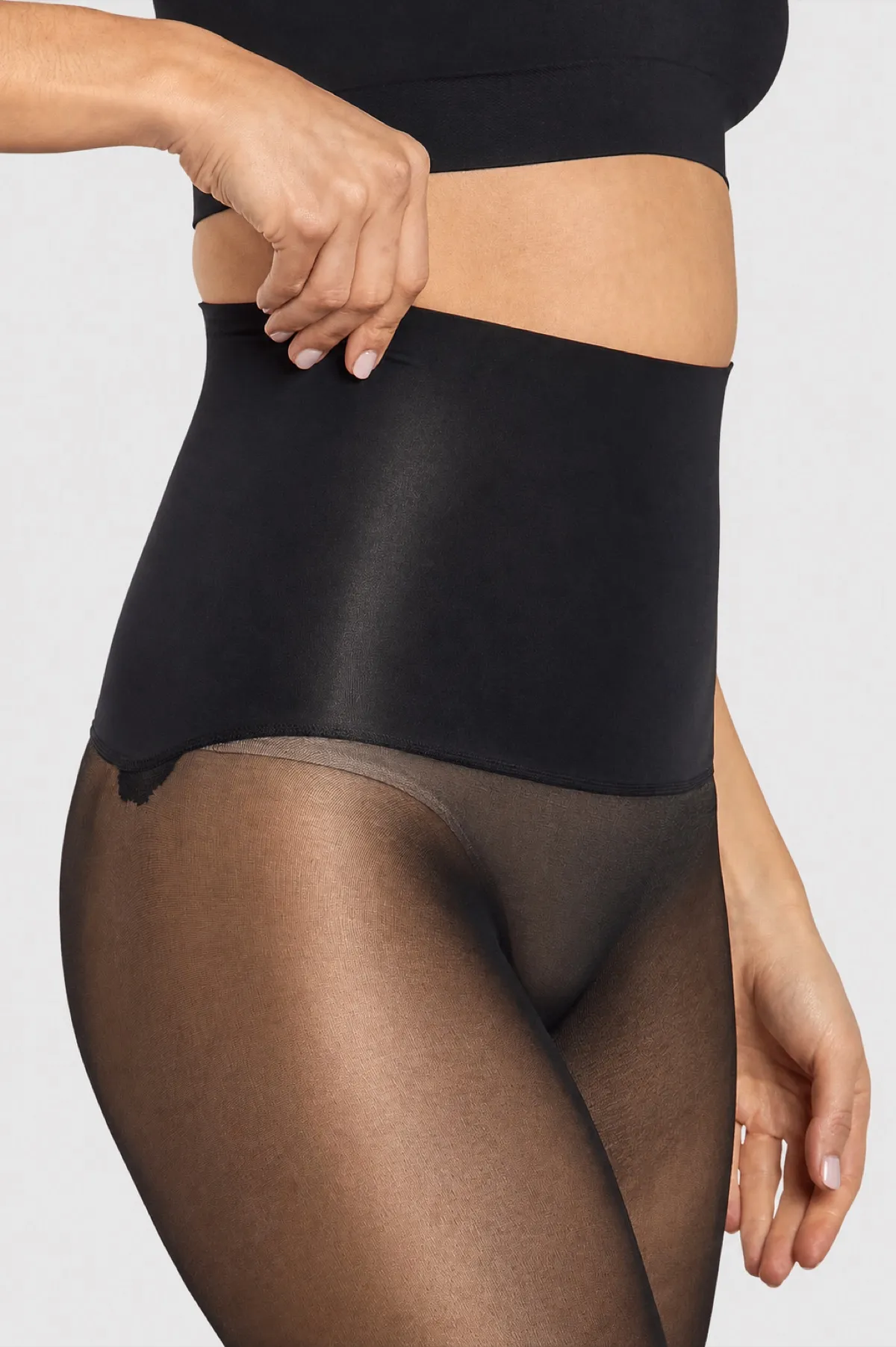 Dettaglio della fascia alta nera del collant Thirty-Five Satin di Heist, tenuta da una mano, con la gamba semi-velata lucida sotto