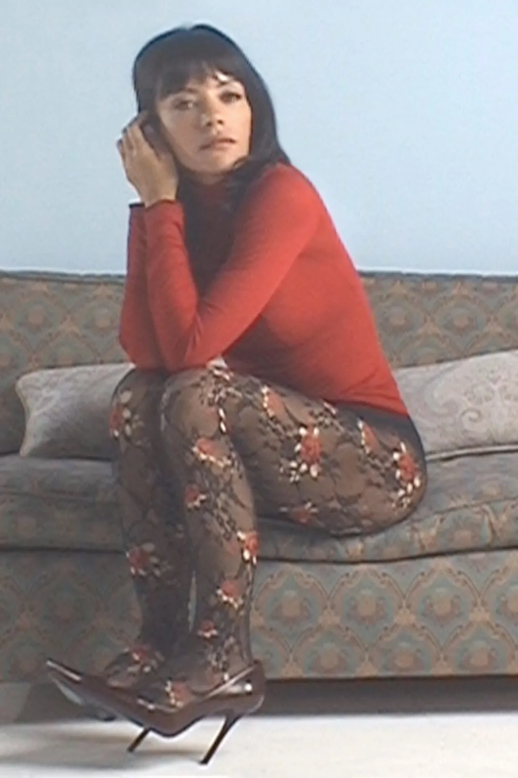 Lily Allen on a sofa in a red roll-neck, floral fishnet tights and black pumps