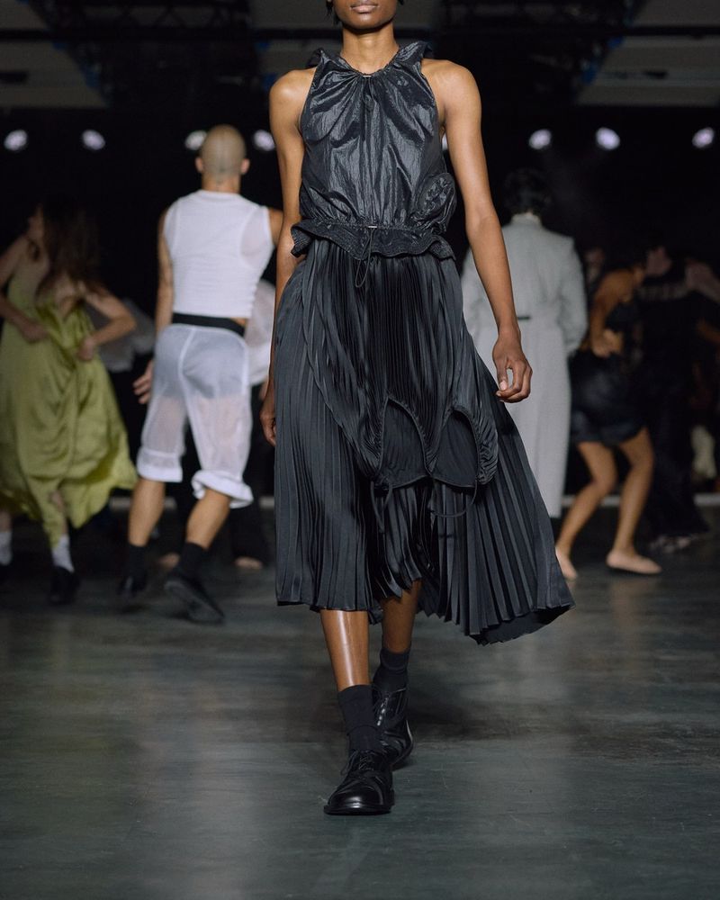 Runway look 21 from Alainpaul spring/summer 2027, a black pleated halterneck dress, black socks and black lace-up shoes