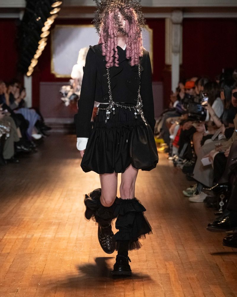 Runway look 22 from Noir Kei Ninomiya spring/summer 2027, a black jacket with chains, a black puffball skirt, black socks with black tulle ruffles on the legs and black studded shoes