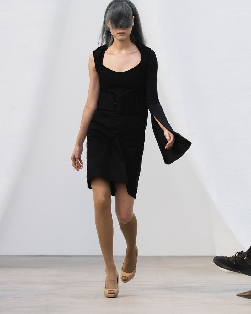 Runway look 22 from De Pino spring/summer 2027, a black dress with one long sleeve, a nude stocking on one leg, a nude knee-high on the other and nude peep-toe pumps