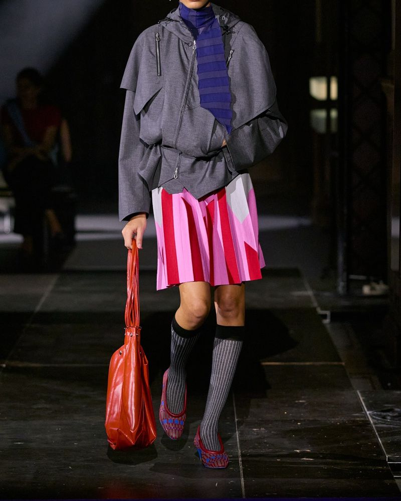 Runway look 24 from Kiko Kostadinov spring/summer 2027, grey ribbed knee socks with a black cuff and woven red and blue pumps under a pink striped pleated skirt and a grey jacket