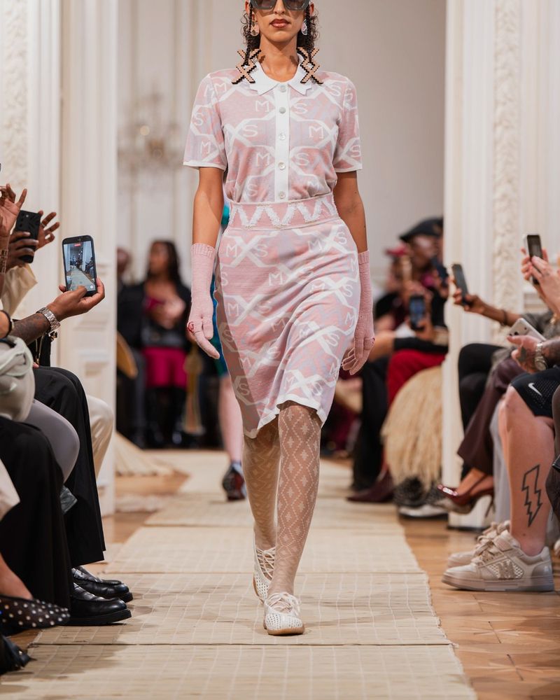 Runway look 27 from Maxhosa Africa spring/summer 2027, a pink knit polo dress with white motifs, white diamond openwork tights and white perforated ballet flats
