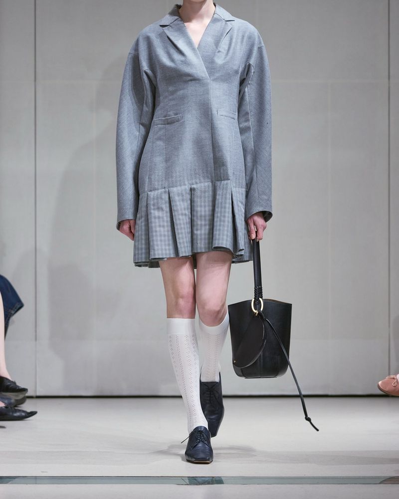Runway look 29 from Mame Kurogouchi spring/summer 2027, a grey check jacket dress with a pleated hem, white openwork knee-highs and navy brogues