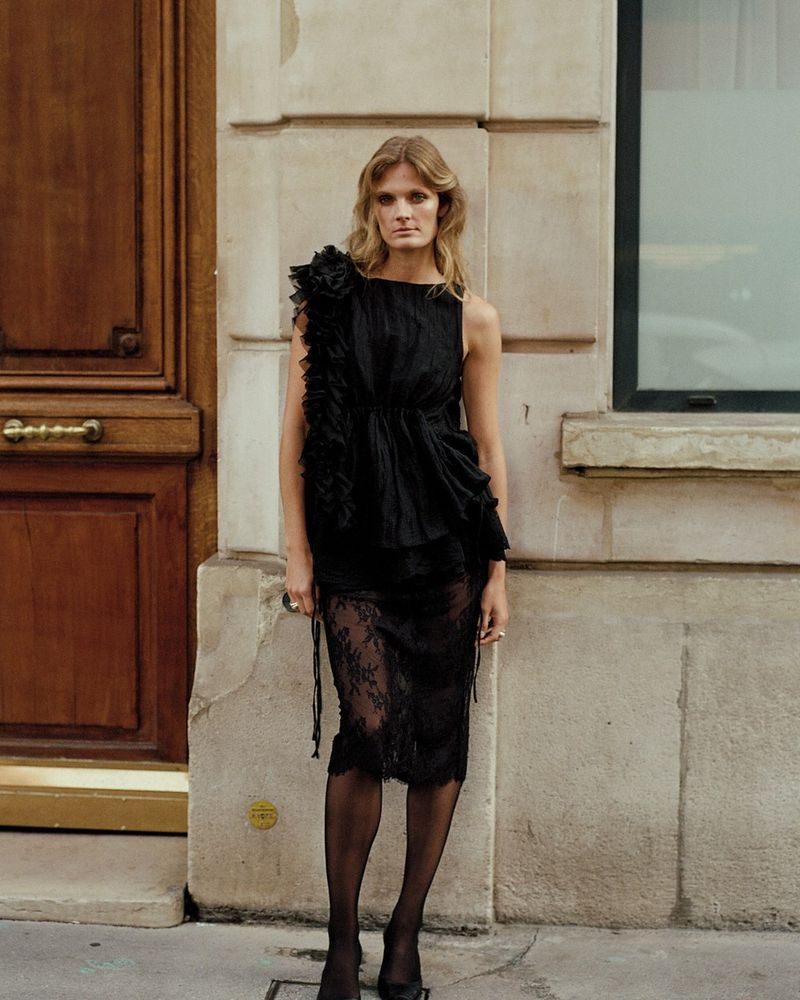 Look 4 from Brock spring/summer 2027, a ruffled black top, a black lace pencil skirt, sheer black tights and black pumps, in front of a wooden door in Paris