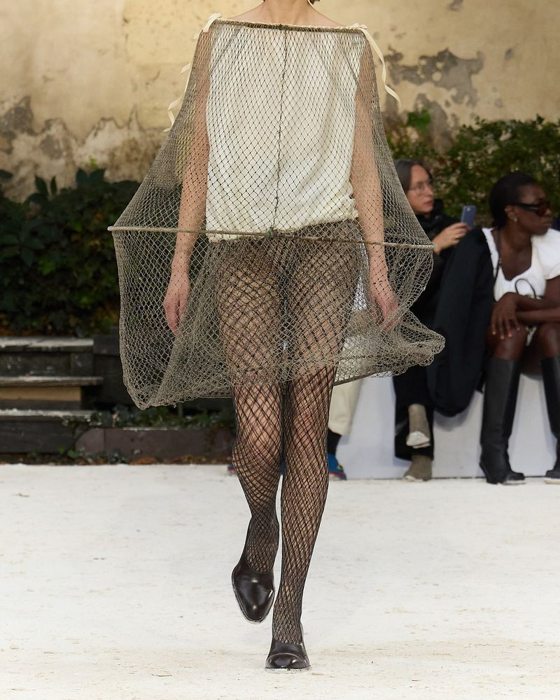 Runway look 42 from Hodakova spring/summer 2027, black fishnet tights and dark pointed flats under a cream top and a fishing net hung from a rope hoop