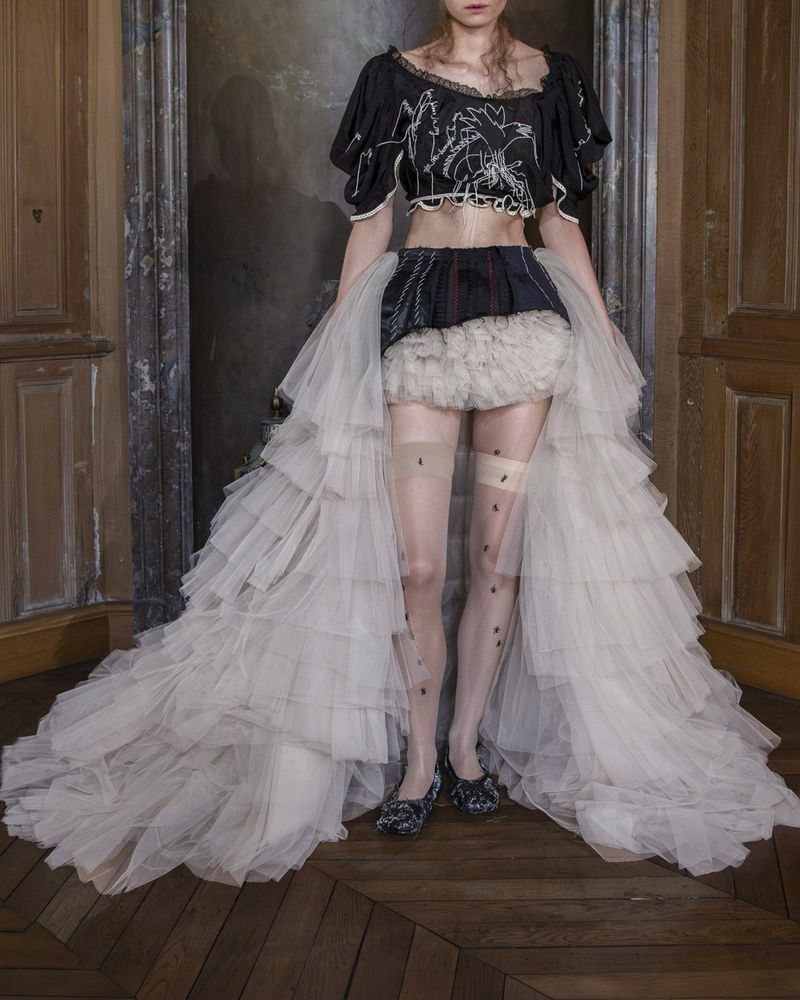 Look 8 from Reverie by Caroline Hu spring/summer 2027, an embroidered black cropped top, a black mini skirt with a tulle ruffle and a long open tulle train, nude over-the-knee stockings with a wide band and small black ants, black ballet flats