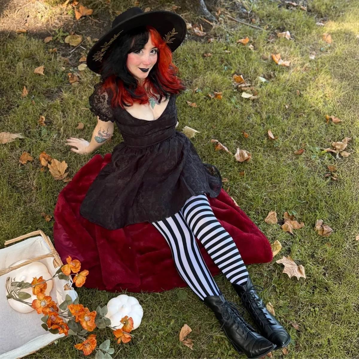 Una donna con i capelli neri e rossi, cappello nero e abito nero di pizzo, seduta su un plaid di velluto rosso nell'erba, con collant a righe bianche e nere verticali su una gamba e orizzontali sull'altra e stivaletti neri allacciati, accanto a zucche bianche e fiori arancioni