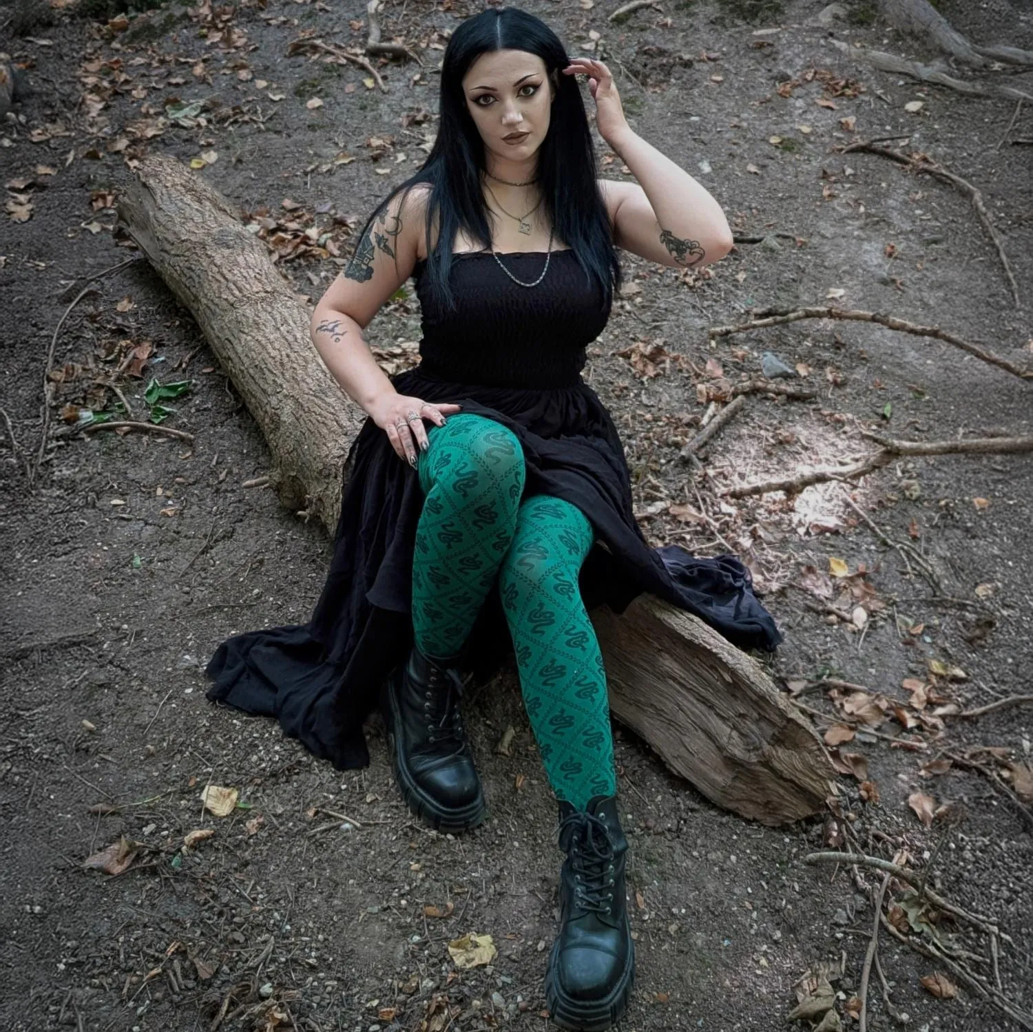 Una donna con lunghi capelli neri seduta su un tronco caduto in un bosco, in abito nero, collant verde scuro con un motivo di piccoli serpenti neri e anfibi neri stringati