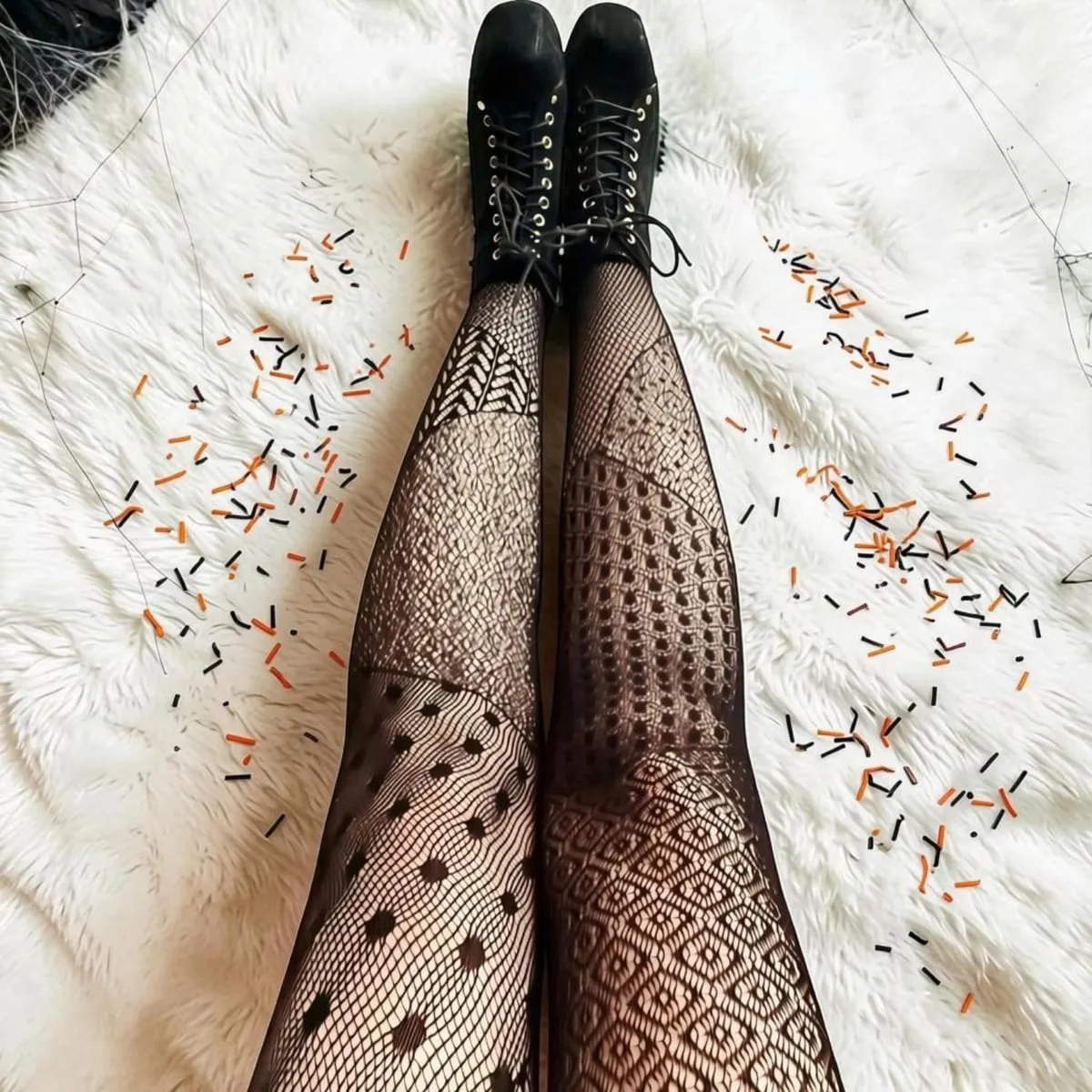 Legs in black fishnet tights with a patchwork of different patterns, polka dots, diamonds and geometric lace, with black lace-up boots, on a white fur rug scattered with orange and black sprinkles