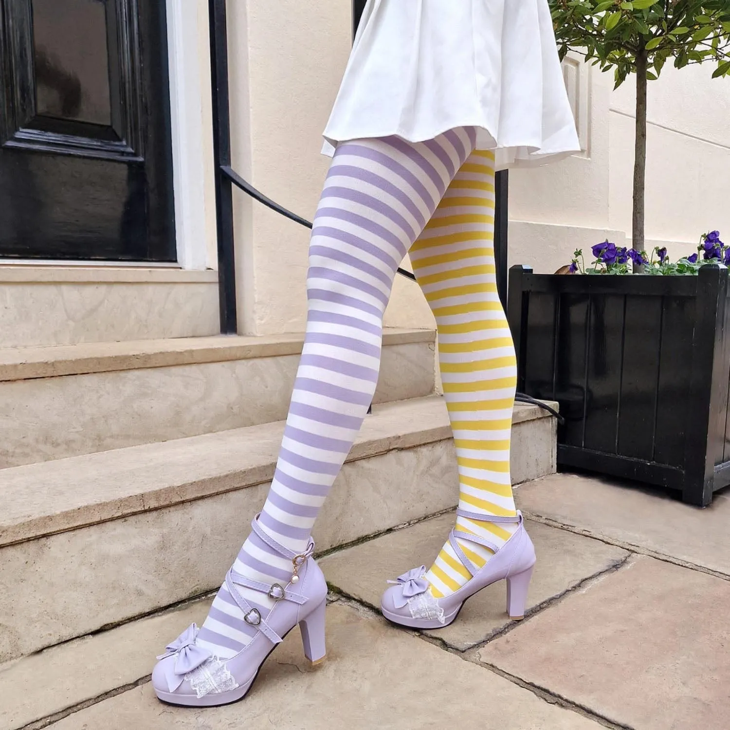 Legs in mismatched striped tights, the left leg lilac and white, the right leg yellow and white, under a short white skirt, with lilac platform Mary Janes with bows, on stone steps