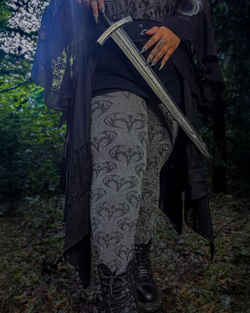 A woman in a long black ragged dress in a dark wood, holding a sword across her hips, in silver grey knitted tights with a repeated dark winged emblem and black lace-up boots