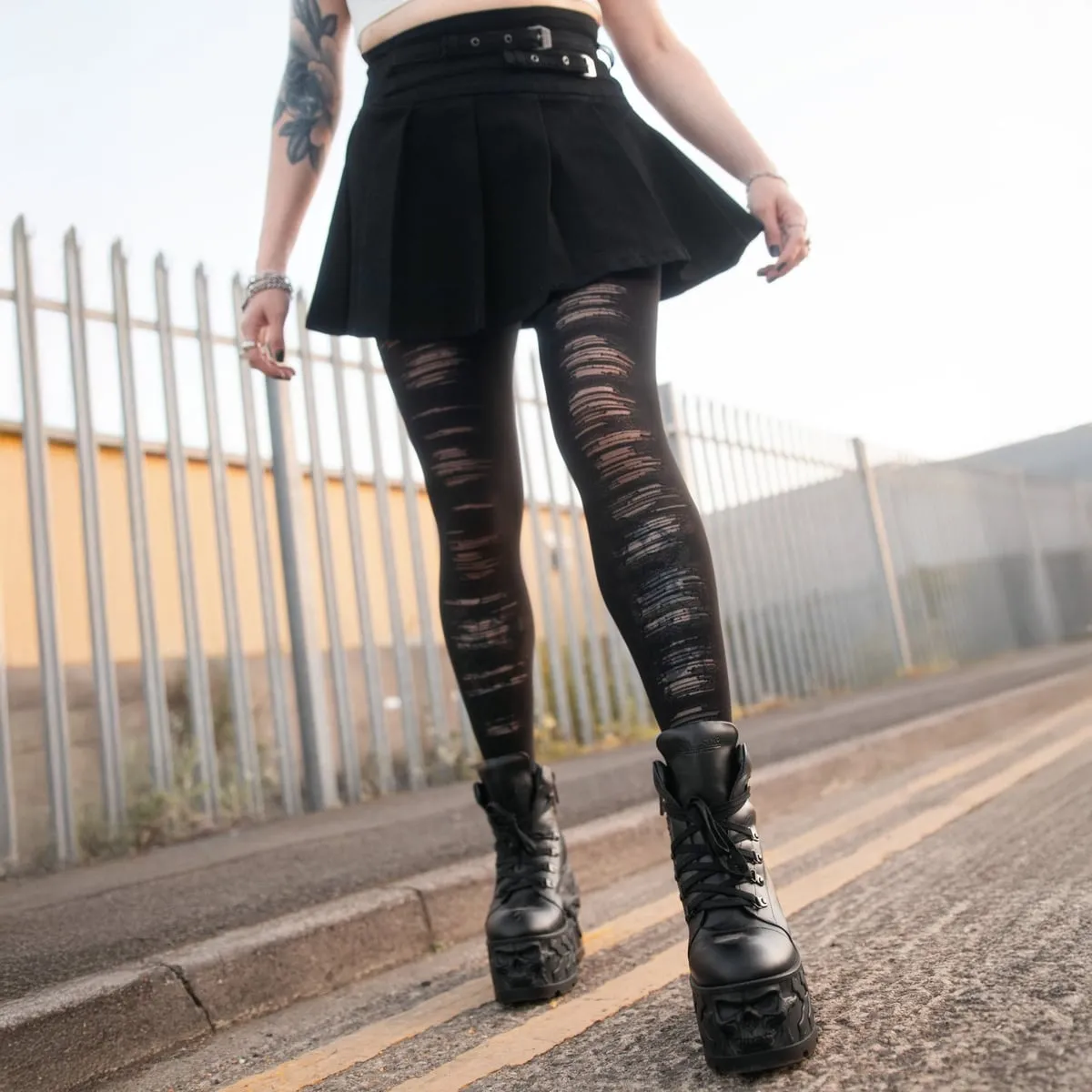Legs in black torn-effect tights under a short black pleated skirt with a belt, with chunky black lace-up boots, on a pavement with yellow lines