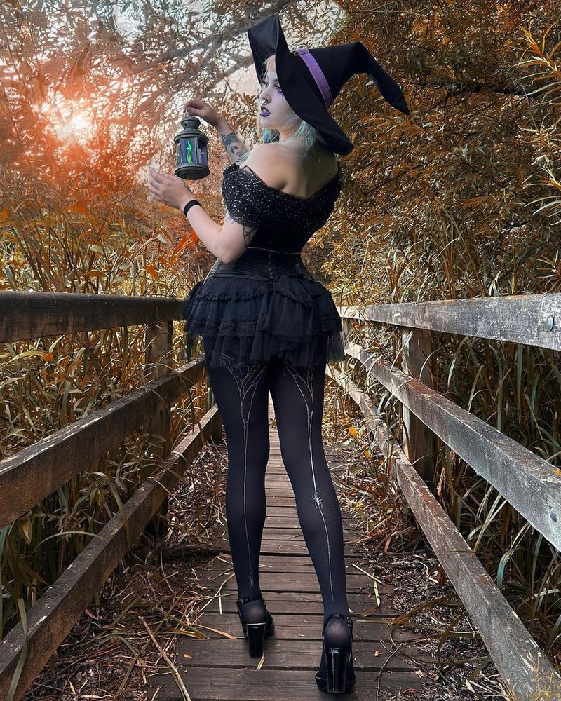 A woman seen from behind on a wooden footbridge among autumn reeds, in a witch's hat, black tutu dress and black opaque tights with a silver glitter cobweb running up the back of each leg, holding a lantern