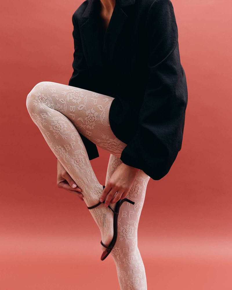 Legs in ivory rose-lace tights with a small BP logo under an oversized black blazer, with thin black strappy sandals, on a coral background