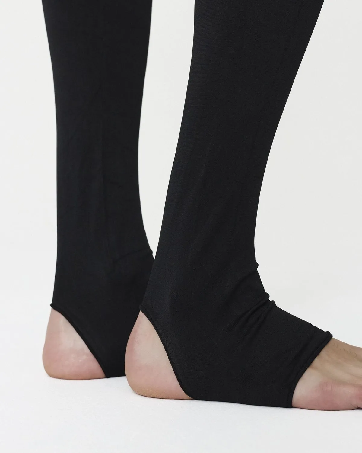 Close-up of black semi-opaque stirrup tights on bare feet: the fabric covers the ankle and instep, the heel and toes are bare
